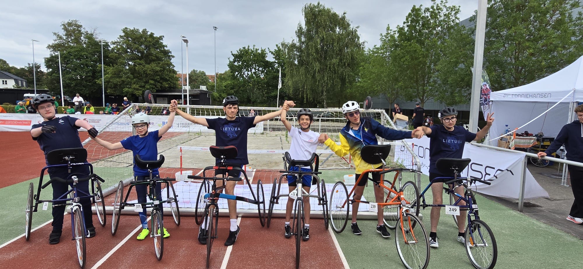 Six young frame runners lined up holding hands on an athletics track at an international frame running event, with a Connie Hansen sponsor tent in the background