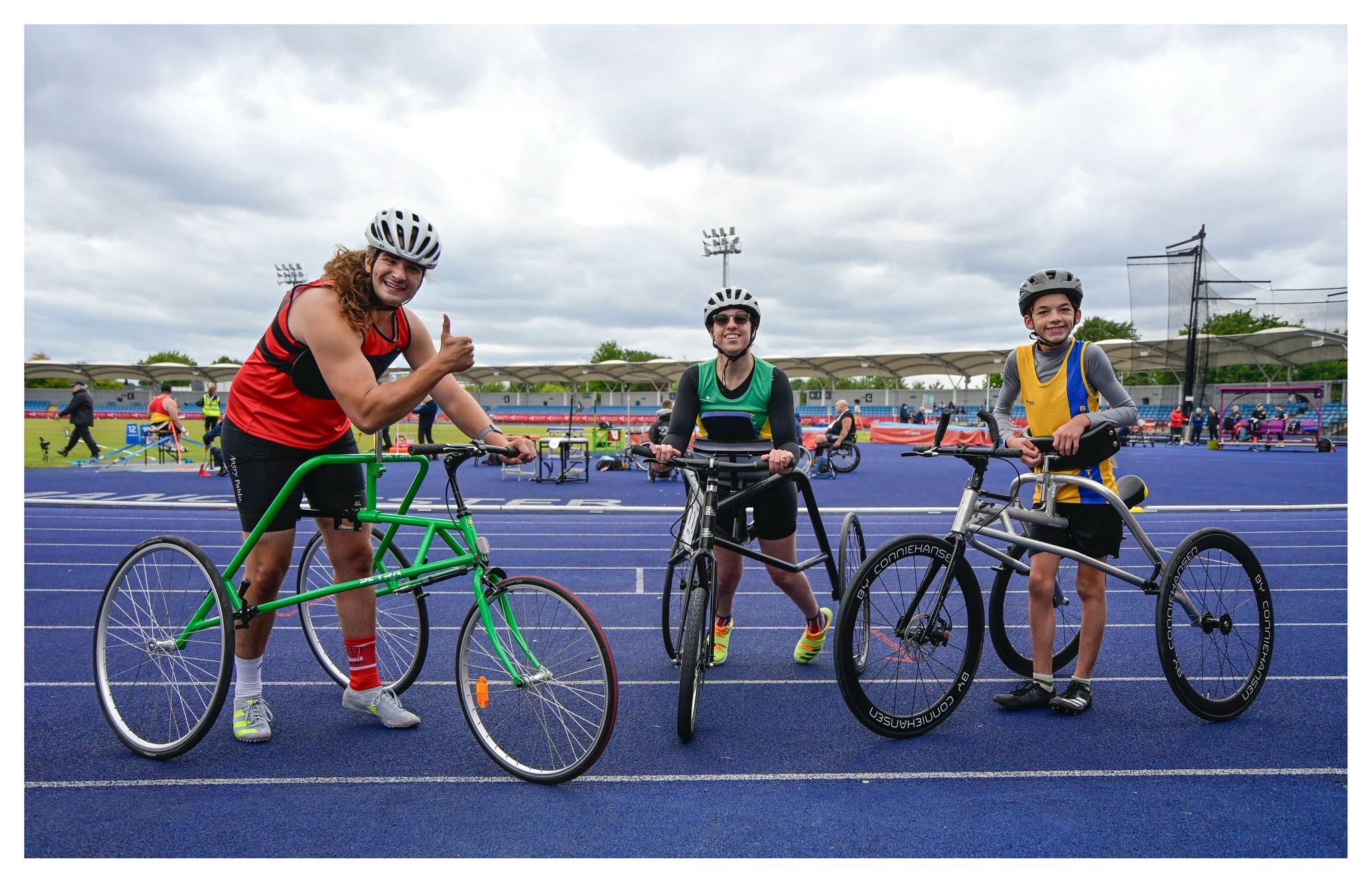 Three athletes standing with their three-wheeled running frames on an athletics track, showing different frame sizes and colours