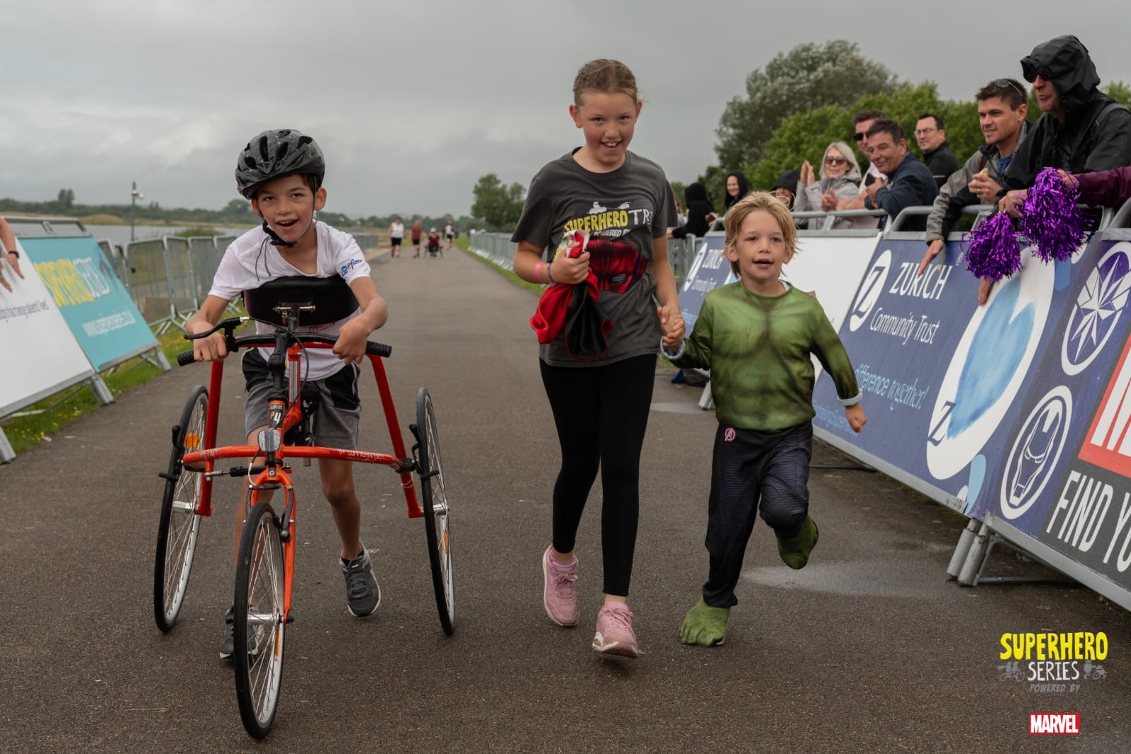 A boy on an orange frame running frame alongside two other children on foot at an outdoor event